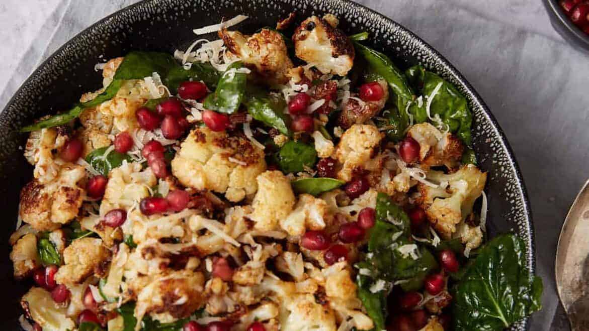 A bowl of roasted cauliflower salad with spinach, shredded cheese, and pomegranate seeds.