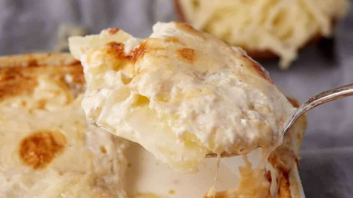 A close-up of a serving spoon lifting a portion of cheesy, creamy potato gratin from a baking dish.