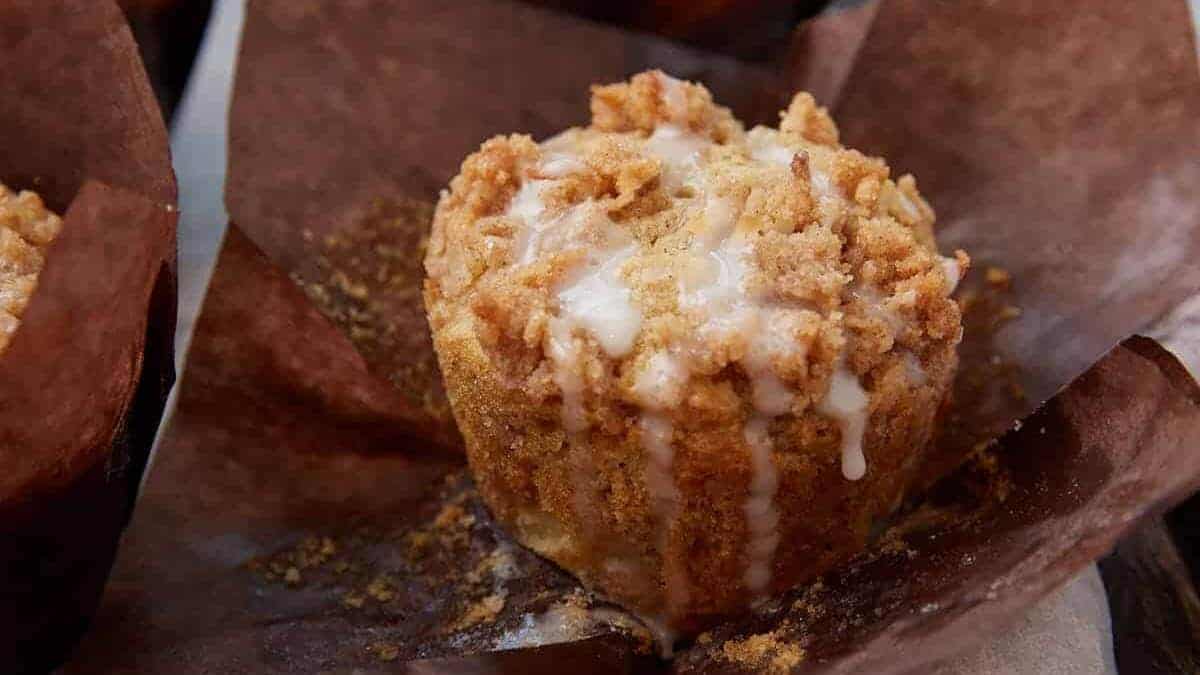 A close-up of a crumb-topped muffin with white icing drizzle, set in a brown paper liner.