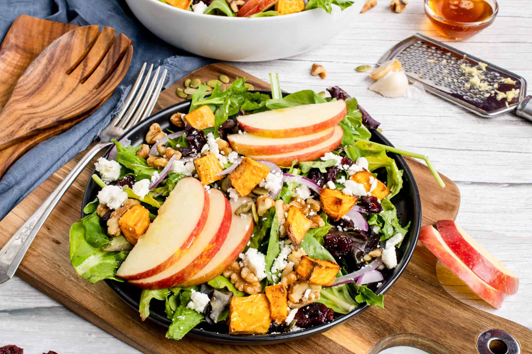 A plate of salad with apple slices, roasted sweet potatoes, mixed greens, feta cheese, red onion, walnuts, and dried cranberries on a wooden table.