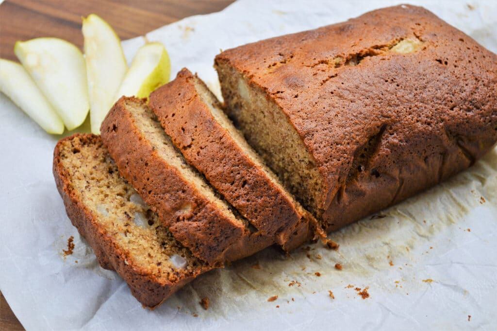 A loaf of pear bread partially sliced on parchment paper, with a few pear slices placed beside it.
