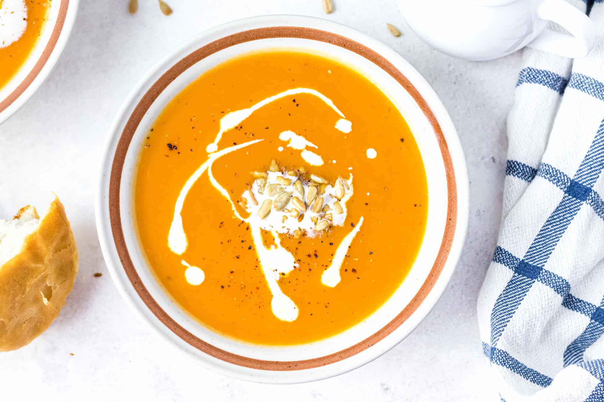 A bowl of orange soup garnished with cream and seeds, next to a piece of bread and a blue checkered napkin.