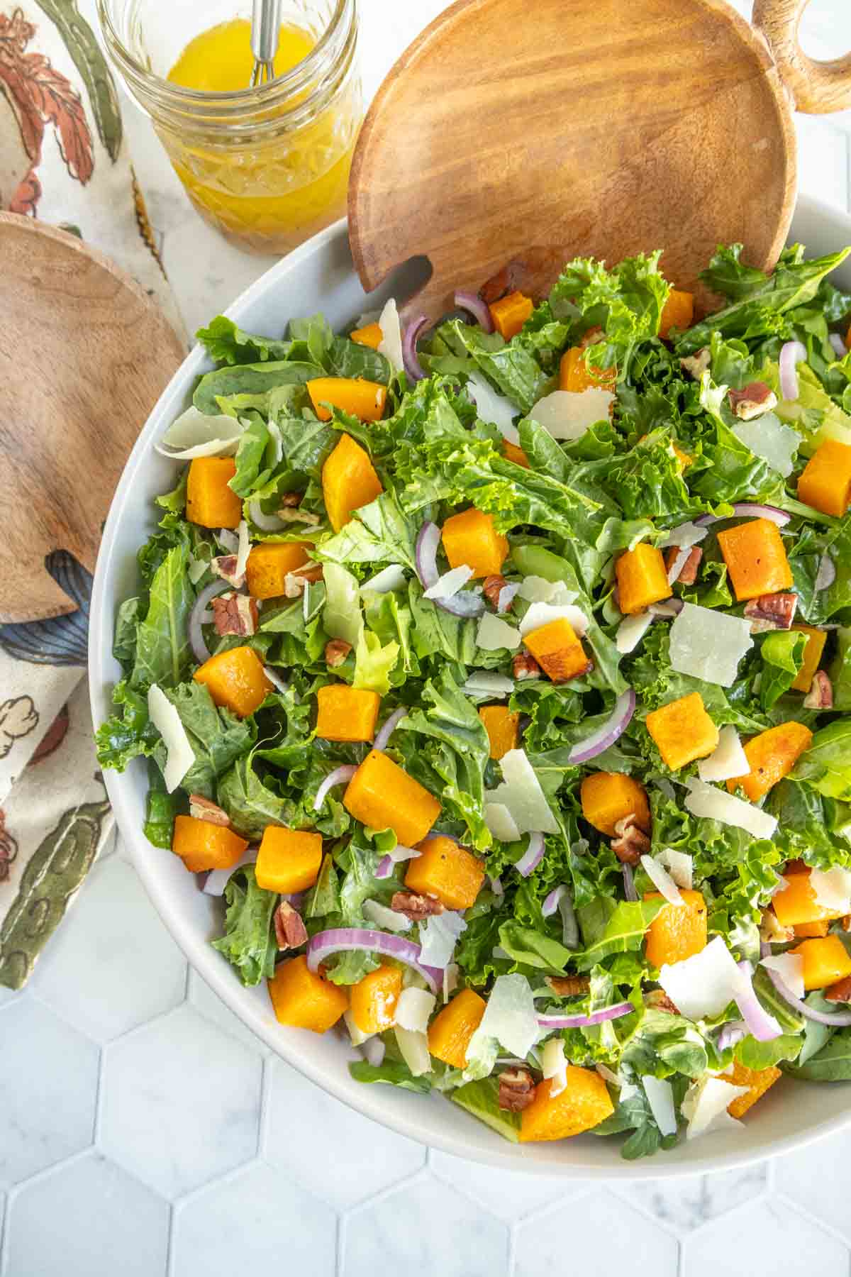 A bowl of mixed green salad with roasted butternut squash, red onion, pecans, and shaved cheese, with wooden salad servers and a jar of dressing nearby.