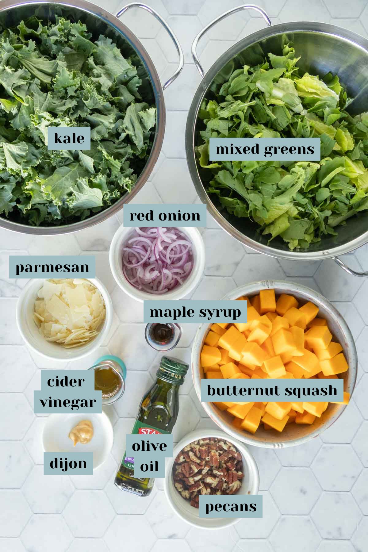 Two bowls contain kale and mixed greens. Surrounding them are bowls and bottles of red onion, parmesan, maple syrup, cider vinegar, dijon, olive oil, butternut squash, and pecans.