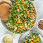 A large bowl of leafy green salad with chunks of butternut squash, red onion, pecans, and shaved cheese, with serving utensils and bowls of toppings on the side.