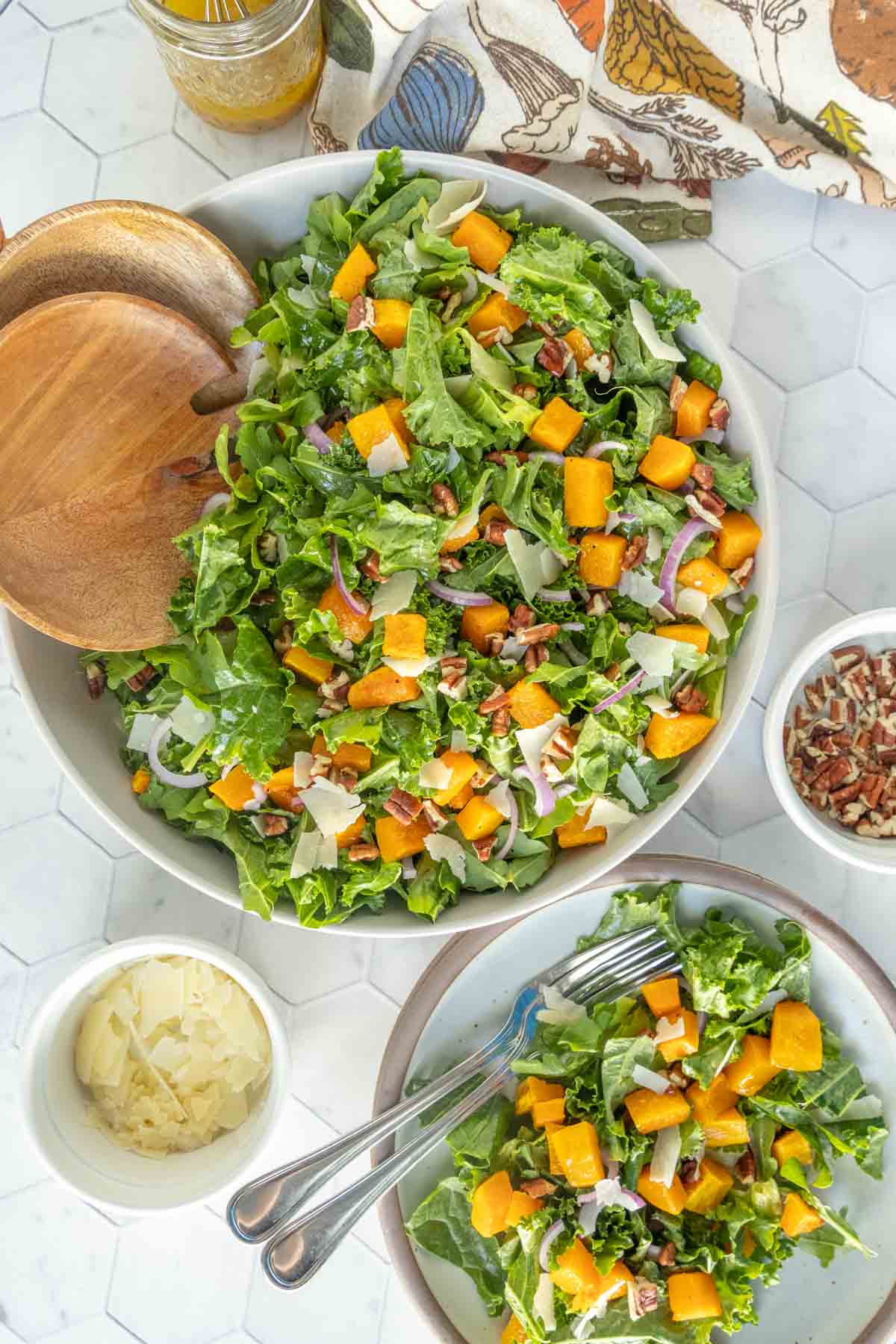 A large bowl of leafy green salad with chunks of butternut squash, red onion, pecans, and shaved cheese, with serving utensils and bowls of toppings on the side.