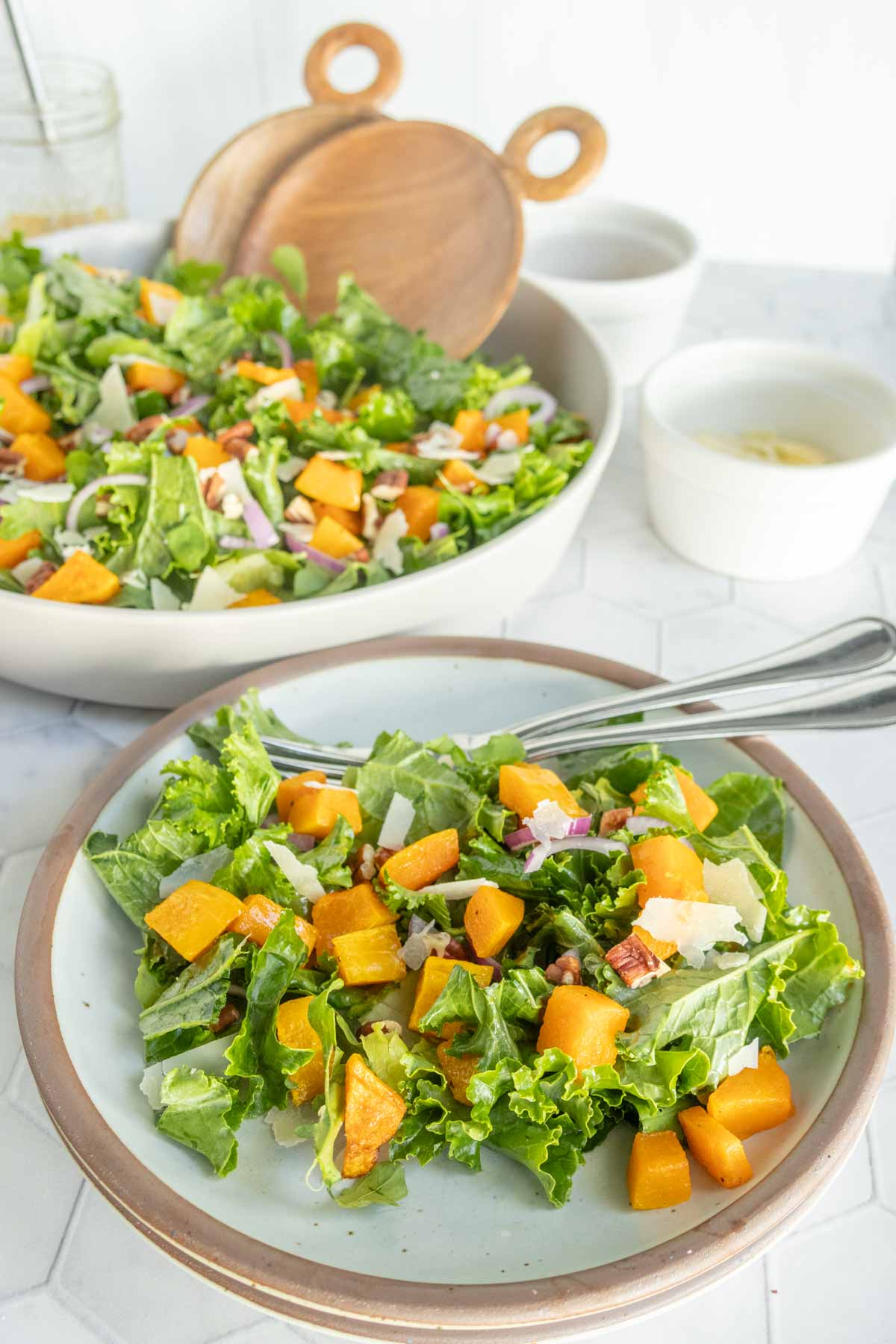A plate of leafy green salad with roasted butternut squash, nuts, red onions, and shaved cheese; a larger bowl of the same salad is in the background.