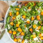 A bowl of butternut squash salad with leafy greens, red onion, shaved cheese, and pecans on a white surface.