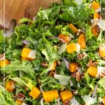 A bowl of butternut squash salad with leafy greens, red onions, pecans, and shaved cheese, shown with a wooden serving spoon.