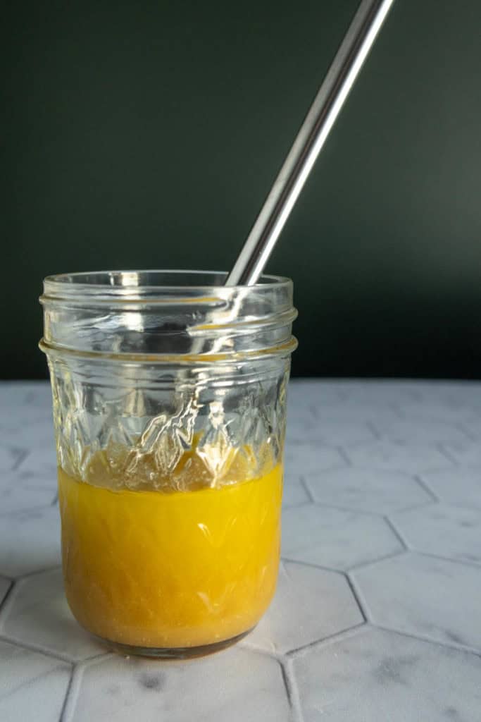 A glass jar containing yellow liquid with a metal spoon inside, placed on a white hexagonal-tiled surface against a dark green background.