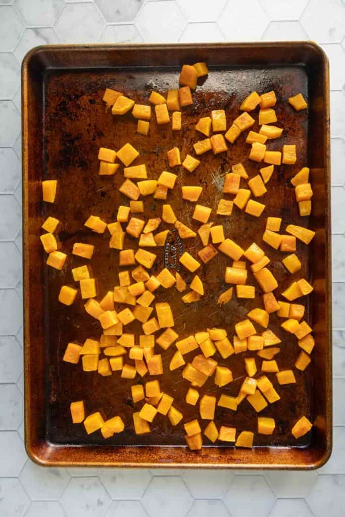Cubed pieces of butternut squash spread evenly on a metal baking sheet, ready for roasting on a white hexagonal tile surface.