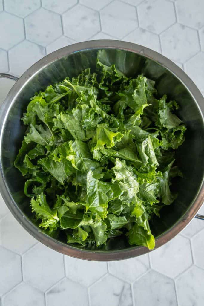 A metal bowl filled with fresh, chopped kale leaves sits on a white hexagonal tile countertop.