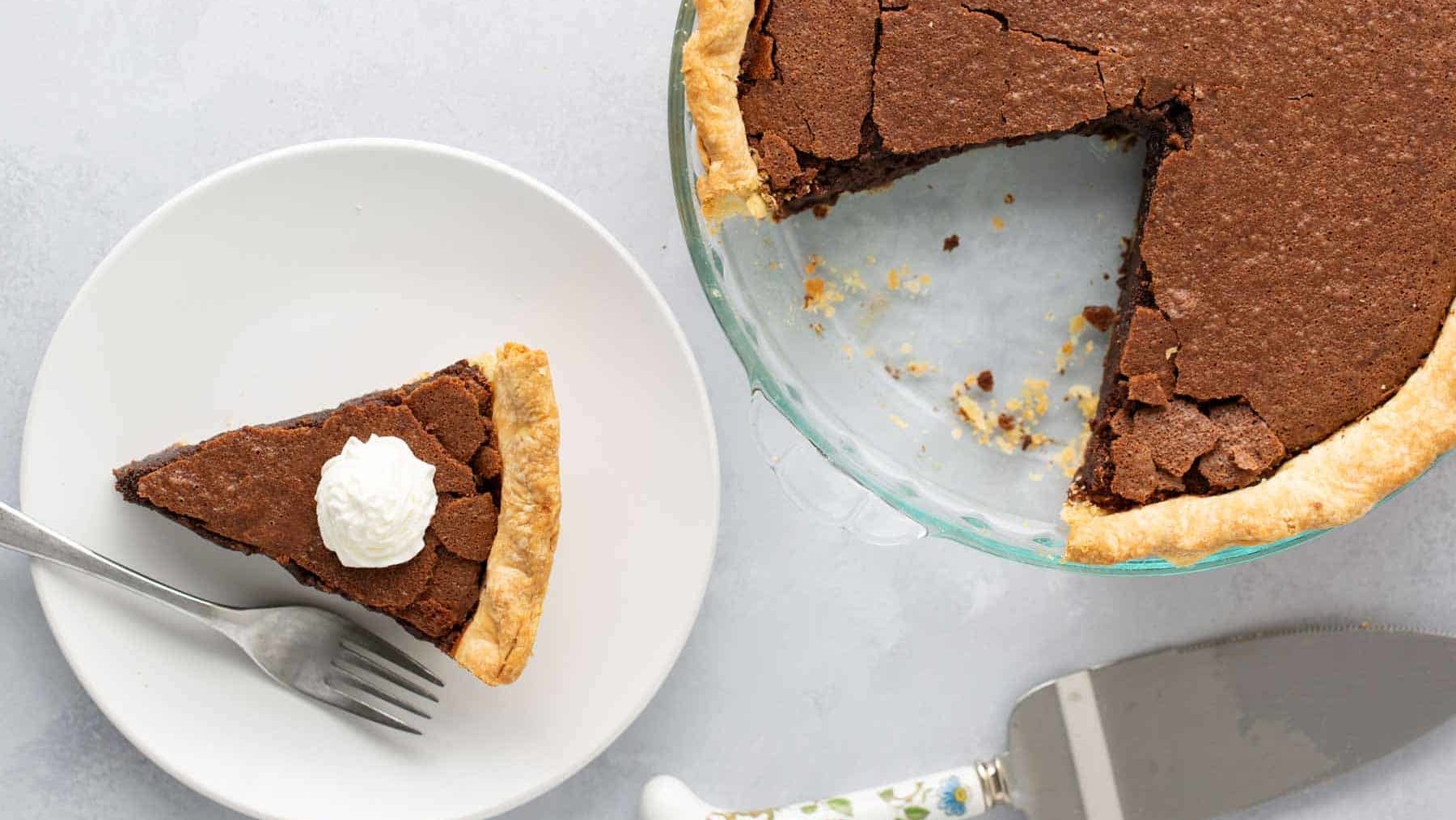 A pie with a missing slice is next to a dessert plate. The plate has a single slice of pie topped with whipped cream, a fork, and a pie server with a floral handle on a light gray surface.