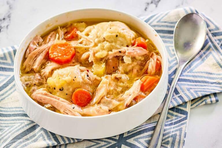 A bowl of chicken and dumplings with shredded chicken, carrots, and broth, placed on a blue-patterned cloth with a spoon beside it.