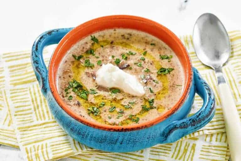 A bowl of creamy soup garnished with chopped herbs, a swirl of oil, and a dollop of sour cream, placed next to a spoon on a patterned cloth.