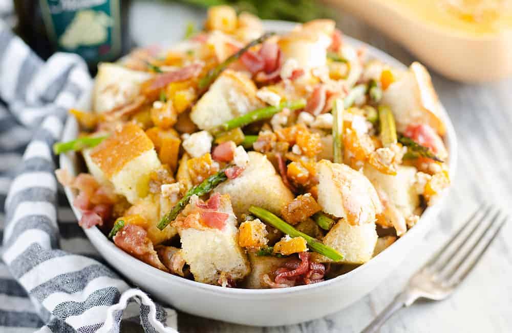 A bowl of panzanella salad with cubed bread, asparagus, butternut squash, prosciutto, and crumbled cheese on a table with a striped napkin and fork.