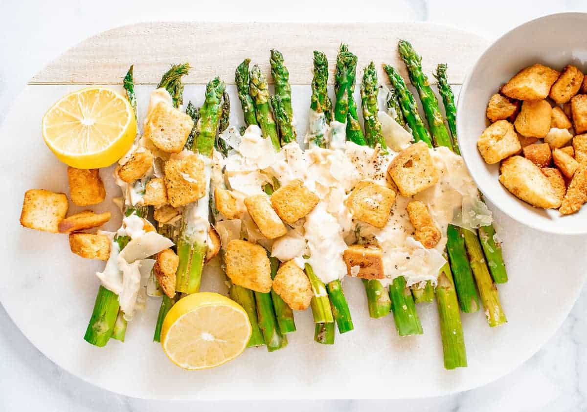 Asparagus spears topped with creamy sauce, croutons, and parmesan, served with lemon halves on a white platter; extra croutons in a bowl nearby.
