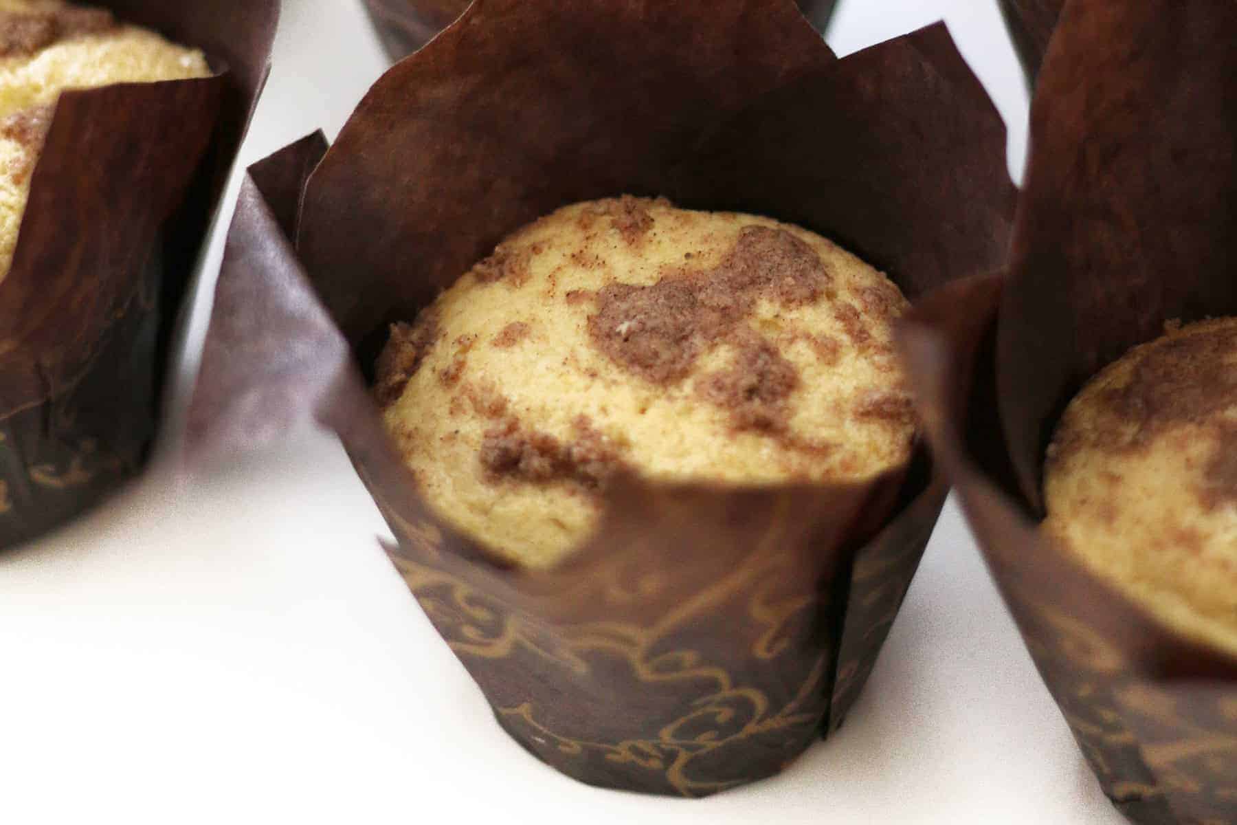 A close-up of muffins with a crumbly topping, each wrapped in brown parchment paper liners on a white surface.