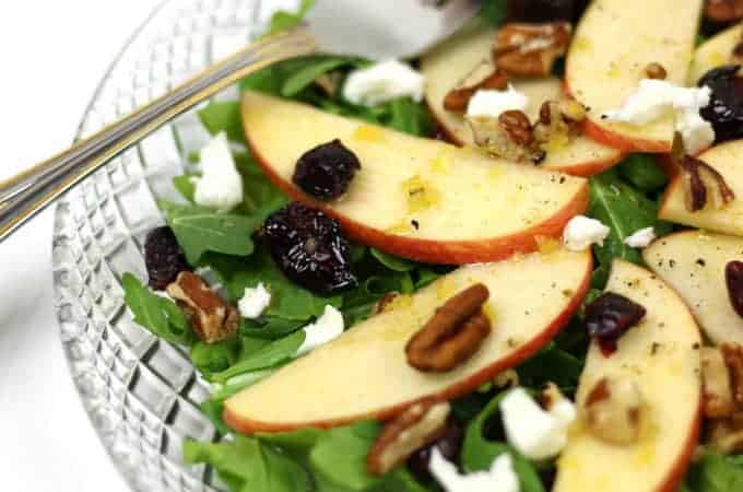 A clear bowl of salad with arugula, sliced apples, pecans, dried cranberries, and crumbled goat cheese, with a fork resting on the edge.