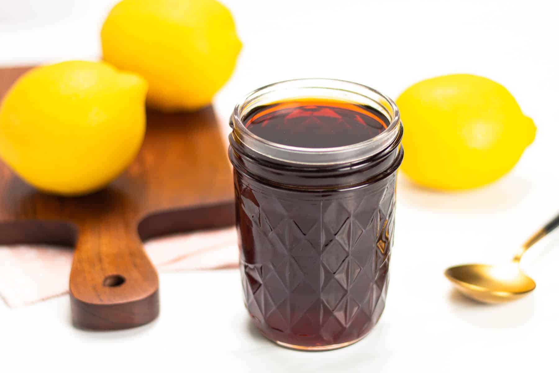 A glass jar filled with dark liquid is placed on a white surface, with three lemons, a wooden board, and a gold spoon in the background.