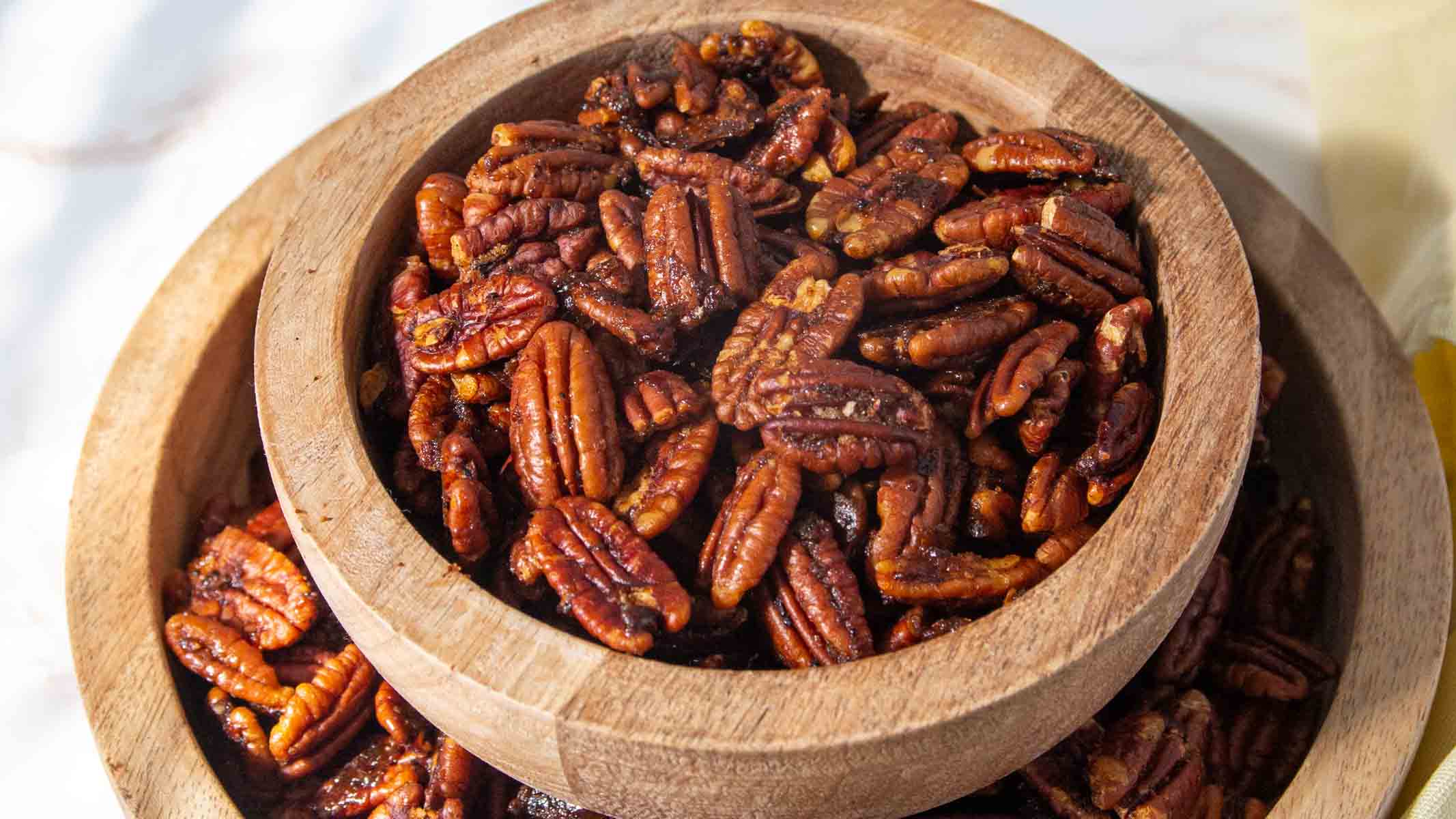A wooden bowl filled with roasted, seasoned pecans sits inside a larger wooden bowl on a light surface.