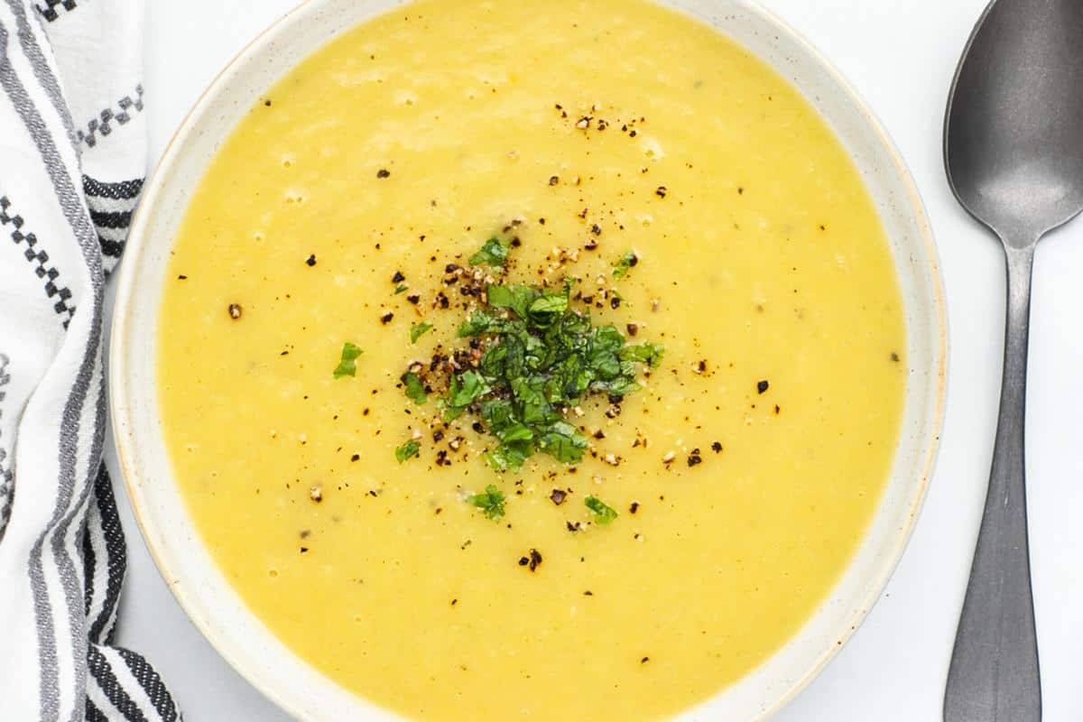 A bowl of creamy yellow soup garnished with chopped herbs and black pepper, placed next to a spoon and a striped cloth napkin.