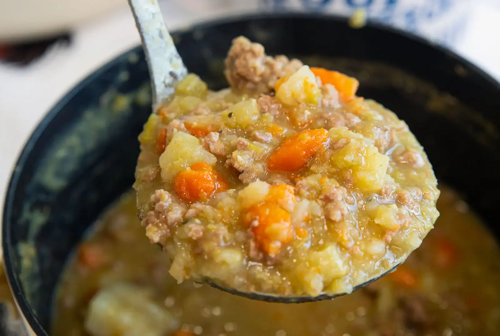 A ladle holds up a thick soup containing ground meat, diced potatoes, carrots, and broth over a pot of the same mixture.