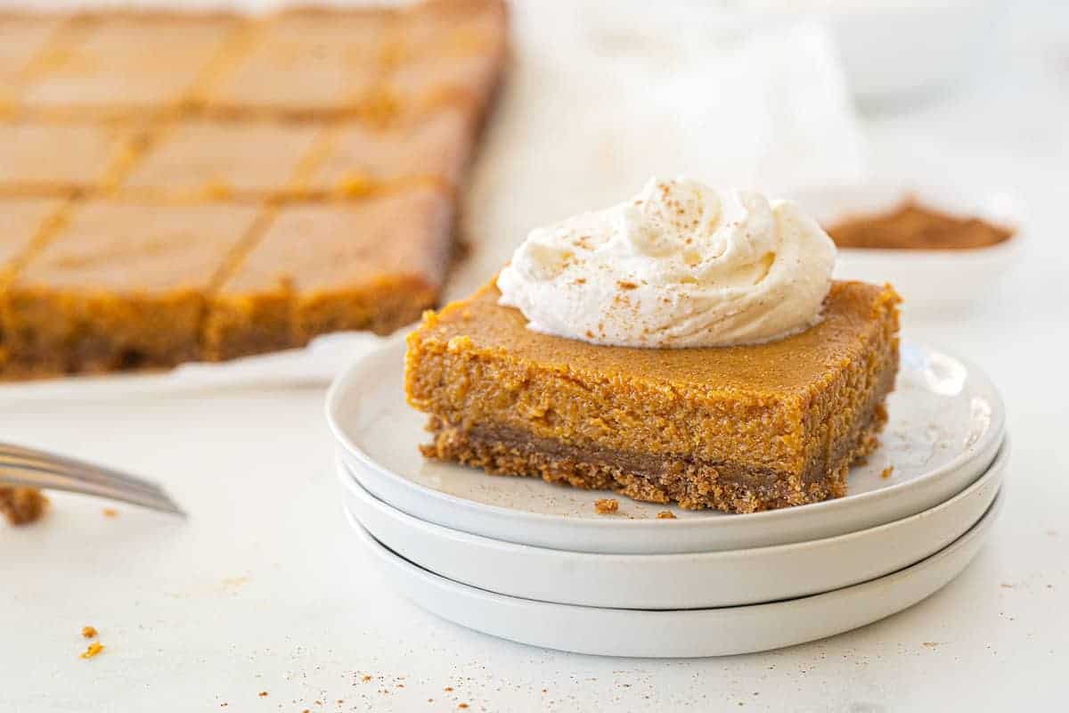 A pumpkin dessert bar with a graham cracker crust is topped with whipped cream on a white plate. The remaining dessert bars are visible in the background.