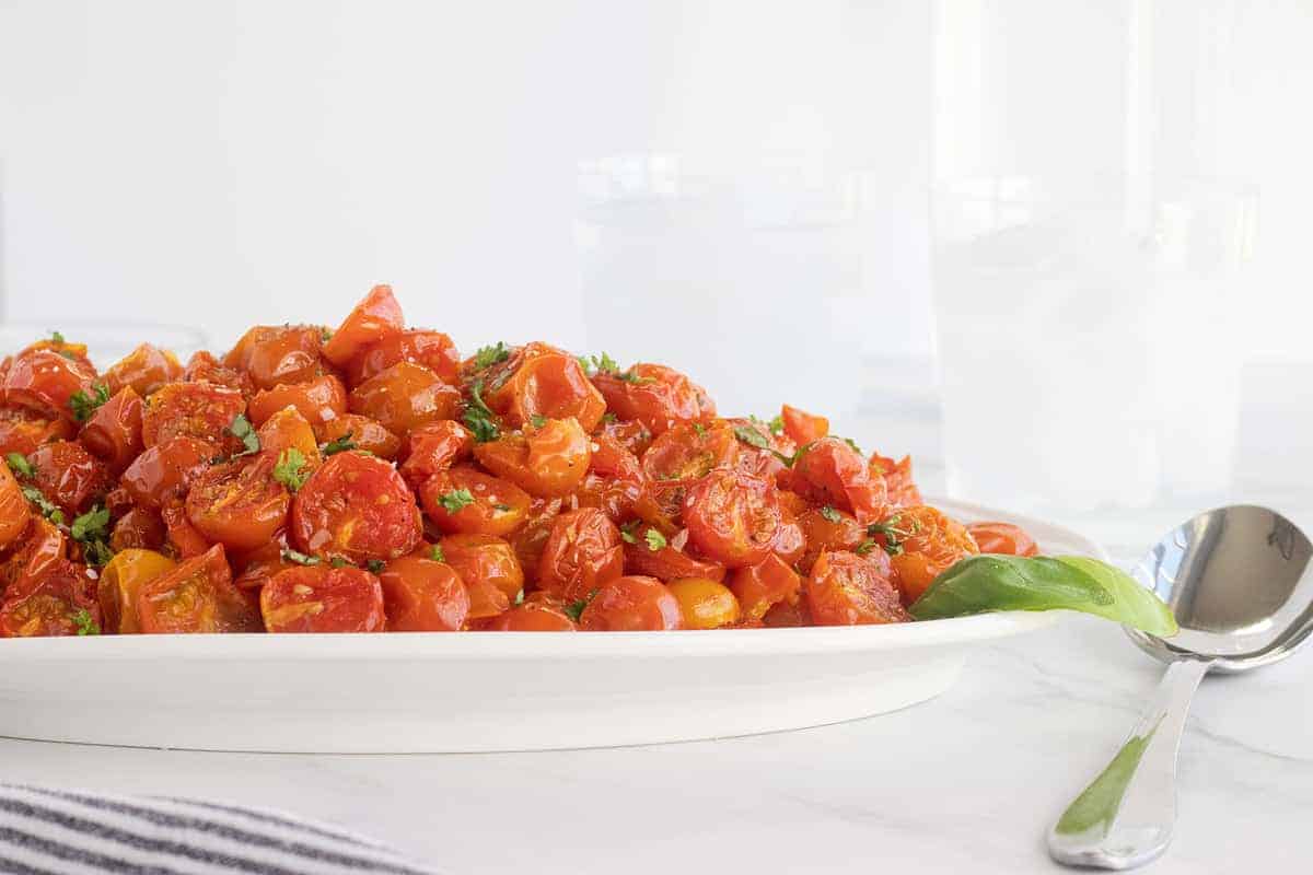 A white platter filled with roasted cherry tomatoes garnished with chopped herbs, placed on a white surface beside a spoon and two glasses of water.