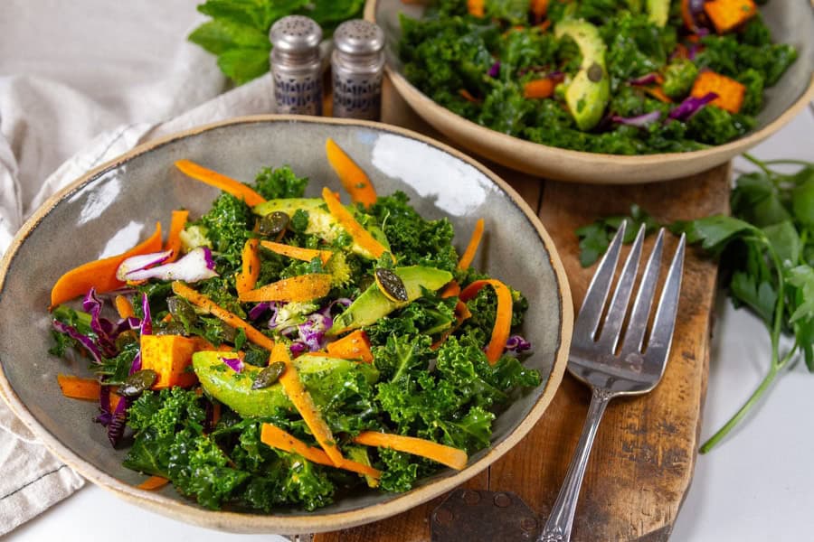 A bowl of kale salad with sliced carrots, avocado, radish, purple cabbage, sweet potato, and pumpkin seeds, with a fork and another salad bowl in the background.