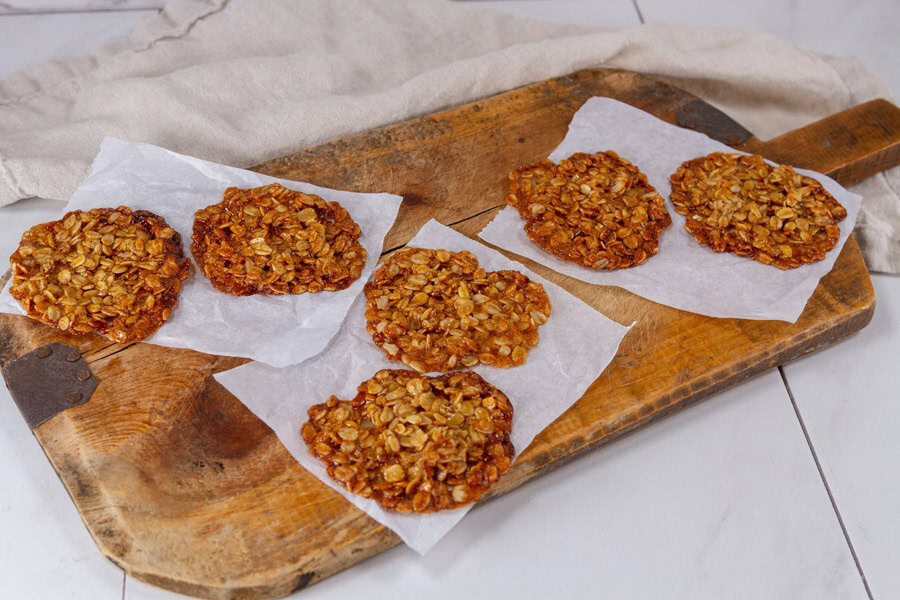 Five oatmeal cookies rest on pieces of parchment paper atop a rustic wooden board, with a beige cloth in the background.