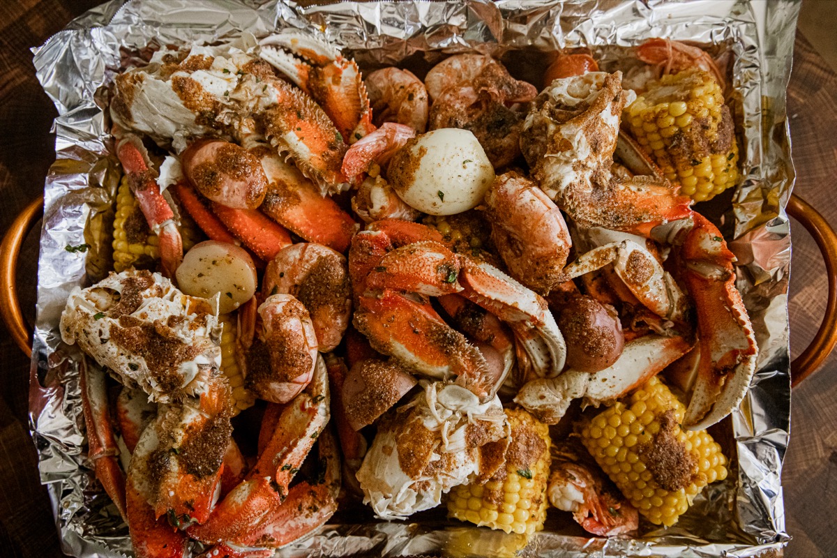 A tray lined with foil holds cooked crab legs, corn on the cob, whole potatoes, and seasoning.