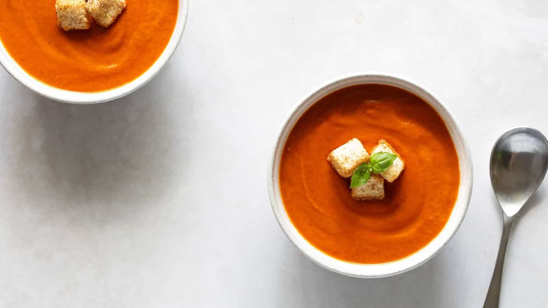 Two bowls of tomato soup topped with croutons and basil leaves, placed on a light-colored surface with a spoon beside one of the bowls.