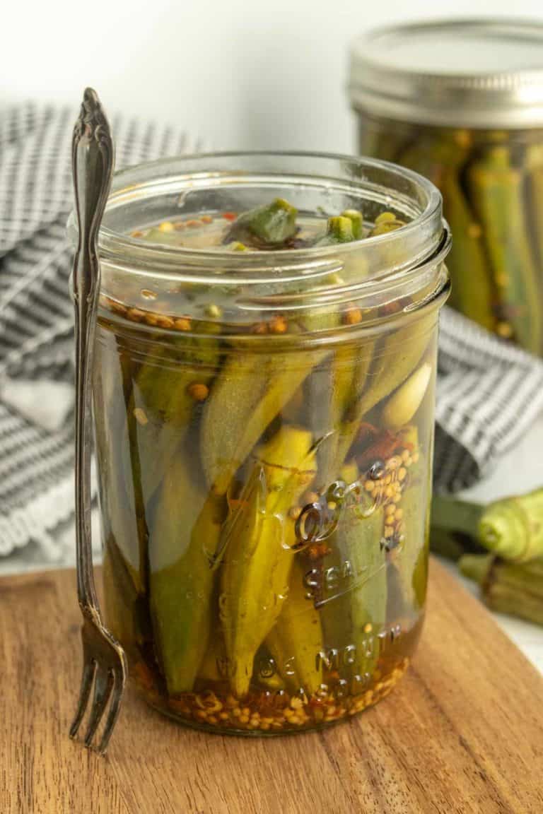 A mason jar filled with pickled okra spears and spices, placed on a wooden surface with a fork resting against the jar. Another jar and a piece of okra are in the background.