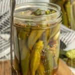 A glass jar filled with pickled okra and spices sits on a wooden board, with a small fork resting against the jar. Another jar is visible in the background.