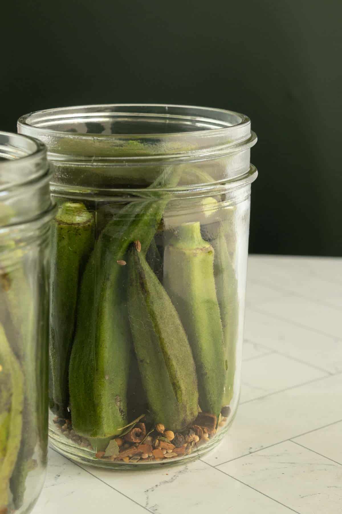 A glass jar filled with whole pickled okra and spices sits on a light-colored countertop against a dark green background.