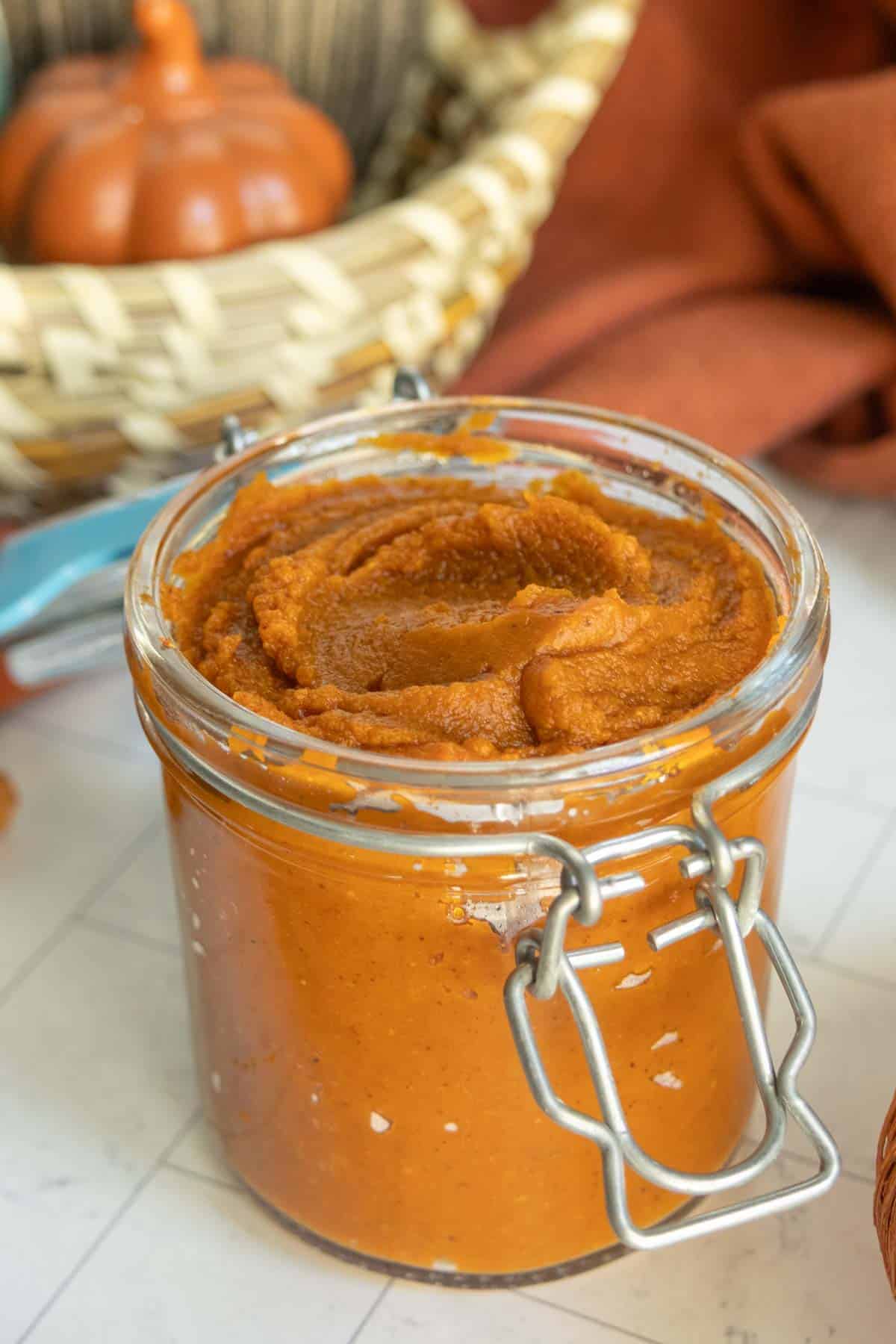 A glass jar filled with thick pumpkin puree sits on a white surface, with a wicker basket and a small decorative pumpkin in the background.