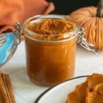 A glass jar filled with pumpkin butter sits on a white surface, next to a plate with bread spread with pumpkin butter and cinnamon sticks.