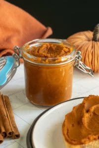 A glass jar filled with pumpkin butter sits on a white surface, next to a plate with bread spread with pumpkin butter and cinnamon sticks.