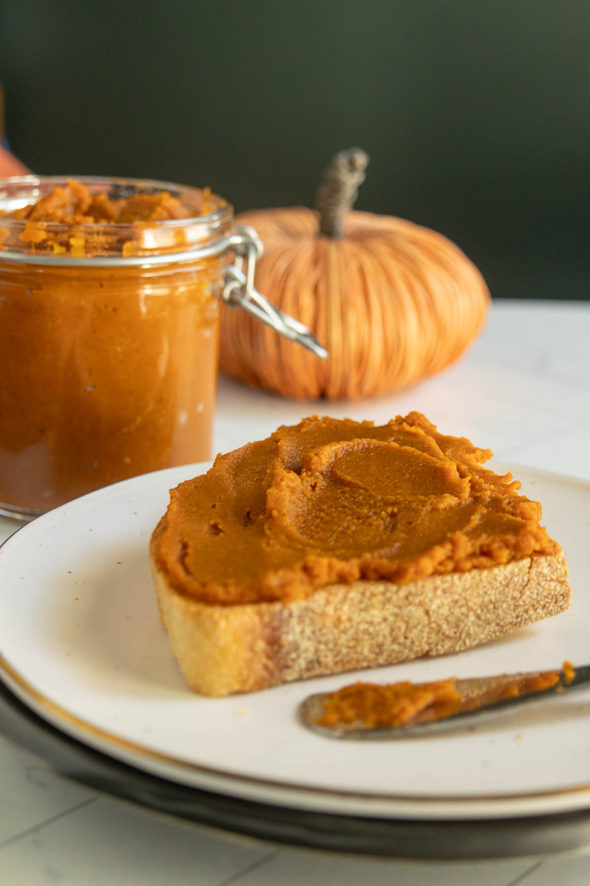 A slice of bread spread with pumpkin butter sits on a plate, with a jar of pumpkin butter and a decorative pumpkin in the background.