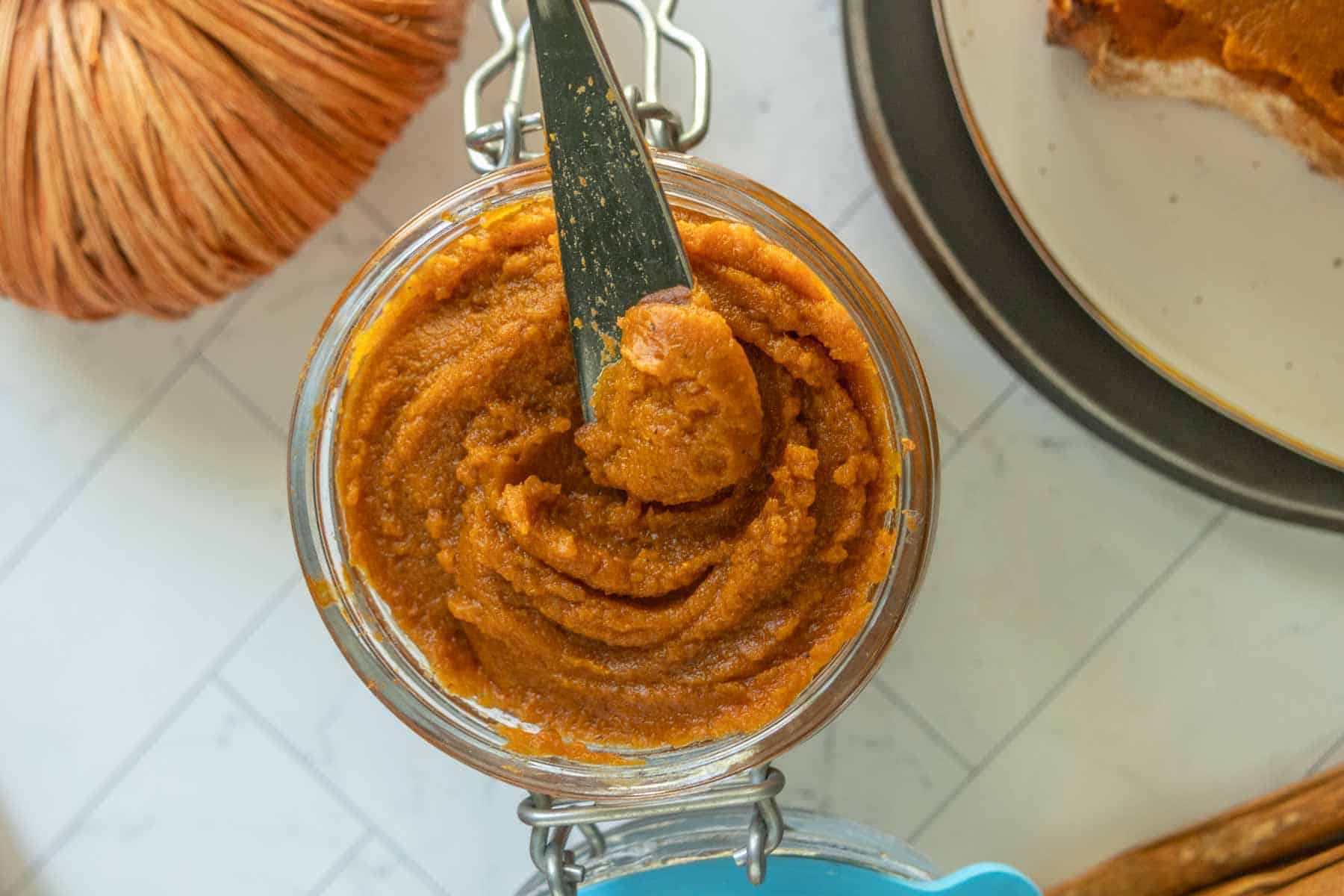 A jar of pumpkin butter with a knife inside, viewed from above, next to a plate with toast and a round decorative object.
