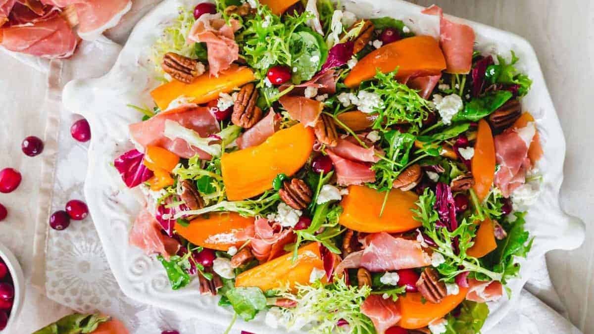 A salad with mixed greens, sliced persimmons, prosciutto, pecans, cranberries, and crumbled cheese on a white platter.