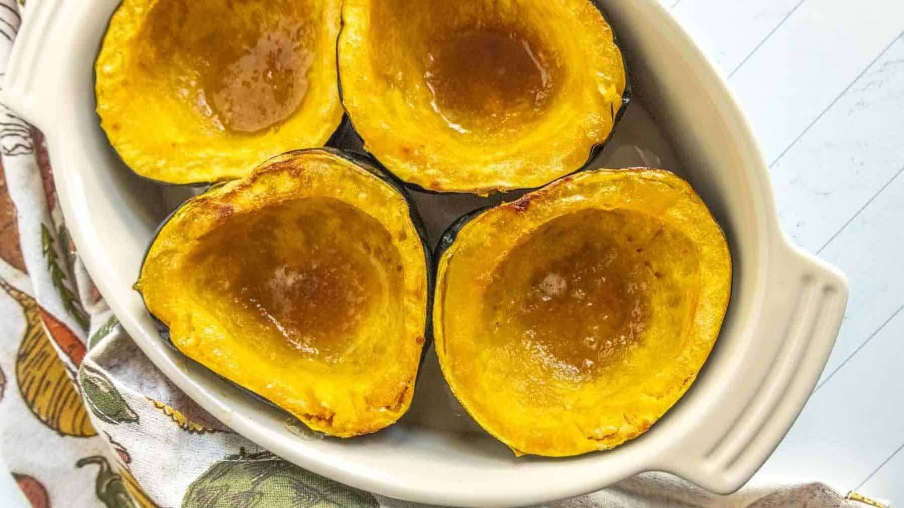 Four baked acorn squash halves with a honey glaze are arranged on a white oval dish. A patterned cloth napkin is placed beside the dish.
