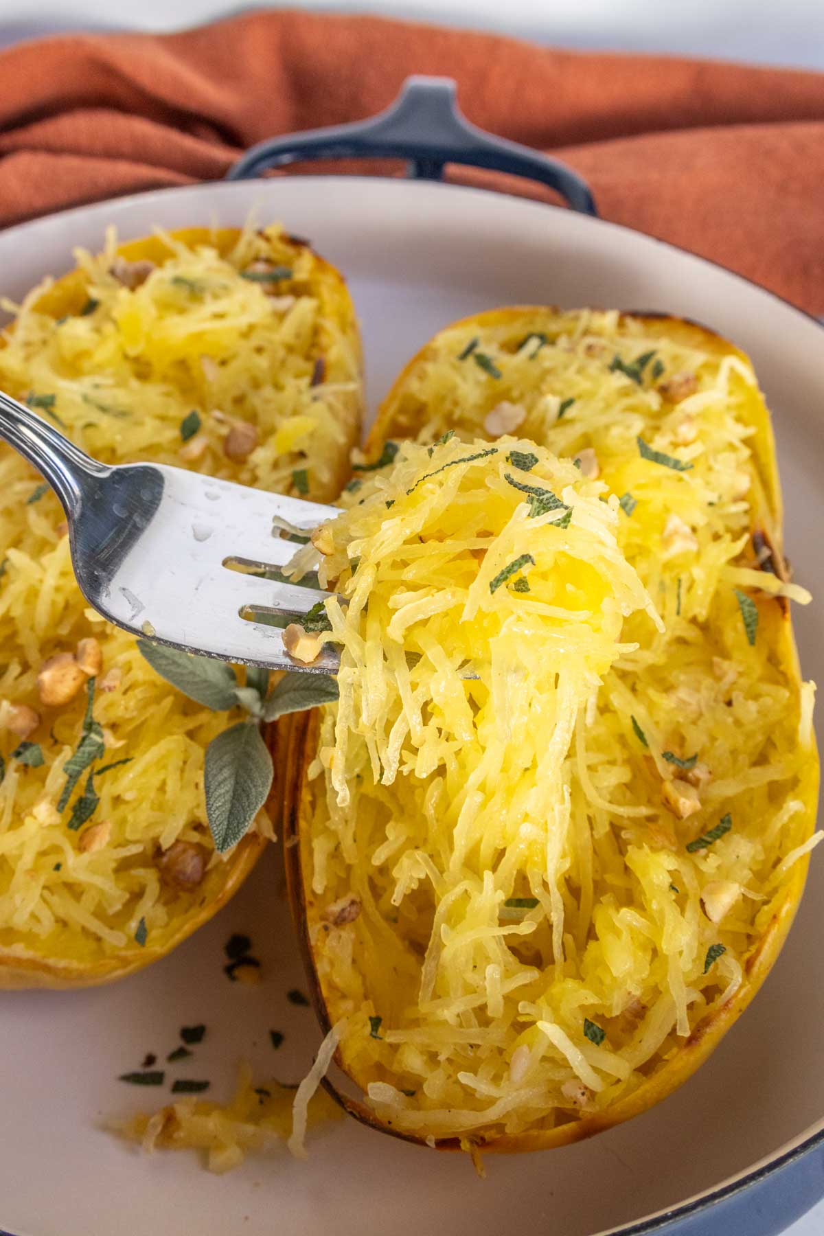 A fork lifting strands of cooked spaghetti squash from a halved squash on a plate, garnished with herbs and small pieces of nuts.