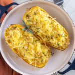 Two baked spaghetti squash halves topped with shredded cheese, herbs, and chopped nuts, served in a round dish on a white surface with a fork and orange napkin nearby.