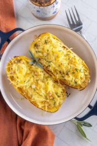 Two baked spaghetti squash halves topped with shredded cheese, herbs, and chopped nuts, served in a round dish on a white surface with a fork and orange napkin nearby.