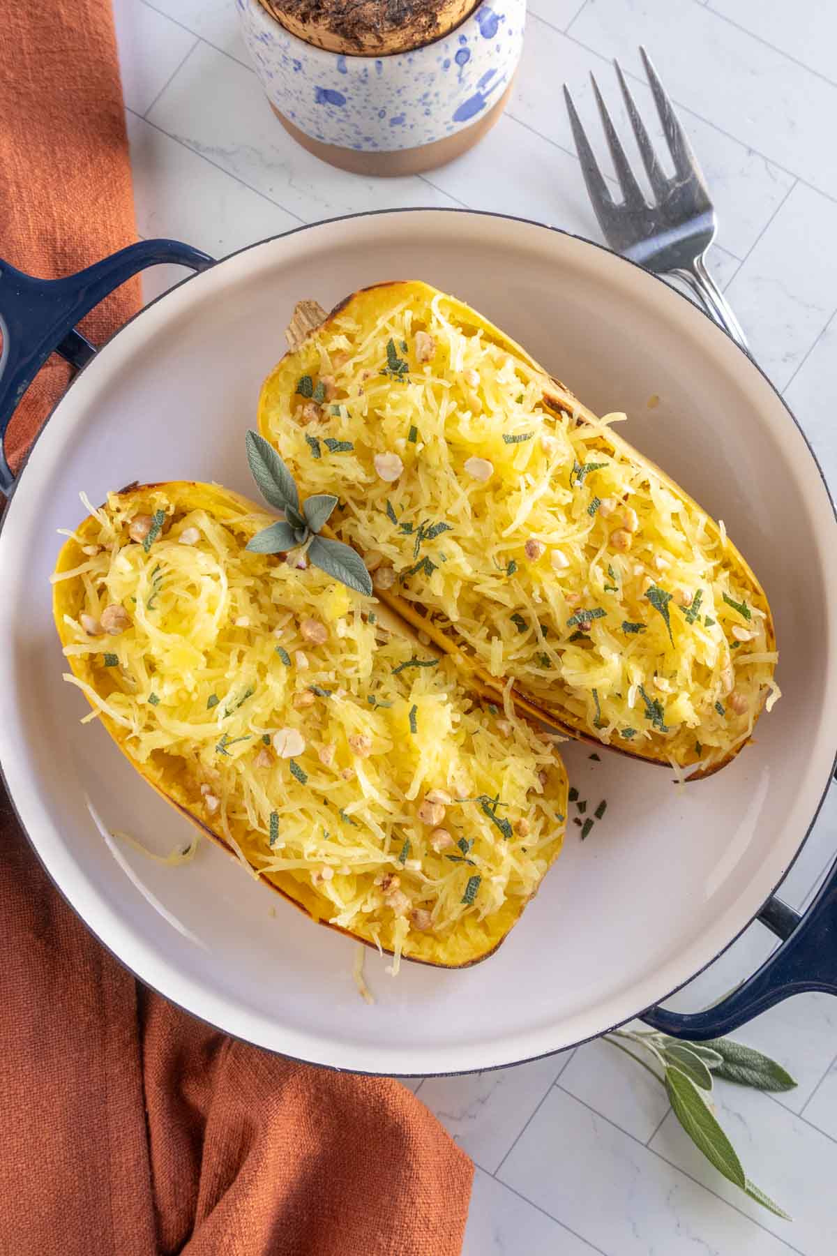 Two baked spaghetti squash halves topped with shredded cheese, herbs, and chopped nuts, served in a round dish on a white surface with a fork and orange napkin nearby.