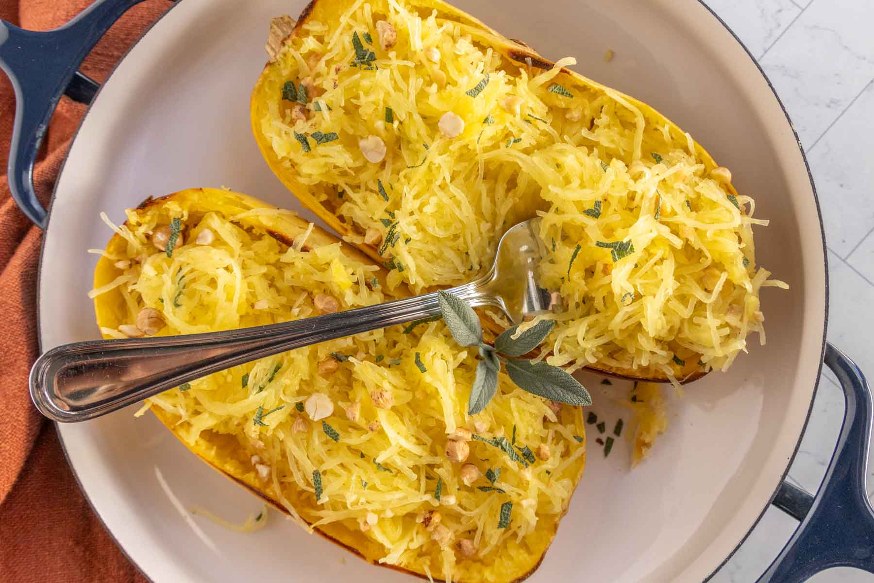 Two halves of cooked spaghetti squash topped with herbs and nuts are served in a white dish with a silver fork.