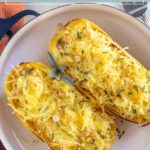 Two halves of roasted spaghetti squash on a white plate, topped with herbs and pine nuts, garnished with sage leaves.