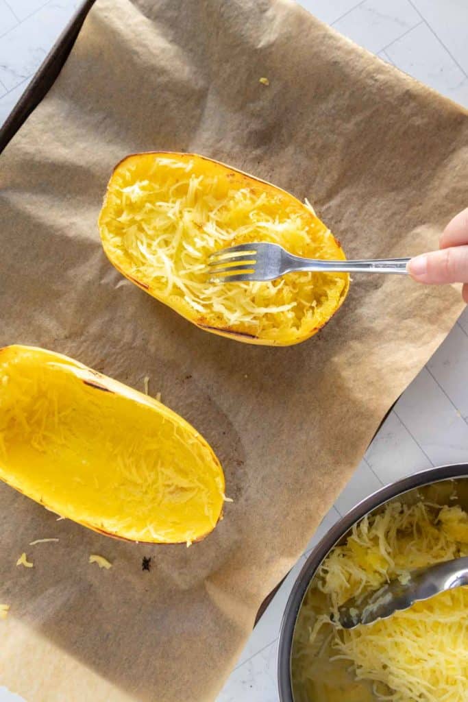 A hand uses a fork to scrape cooked spaghetti squash on a parchment-lined baking sheet, forming noodle-like strands.